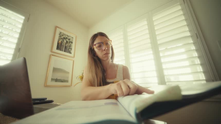 Teenage girl sitting at a desk with a laptop computer as she turns pages of a book