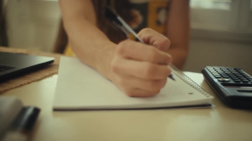 A woman wearing orange glasses and a crochet top flips a page in her notebook and writes with a black pen while sitting at a desk with a laptop and calculator in a sunlit room with white window shutters