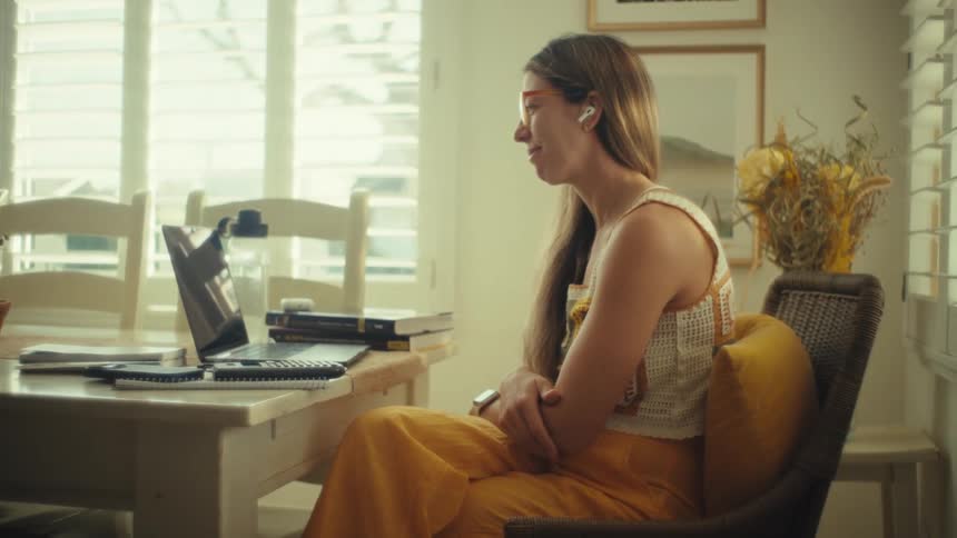 A woman wearing orange glasses and a crochet top sits at a dining table giving a thumbs up during a video call on her laptop while sunlight filters through white window shutters in a bright room