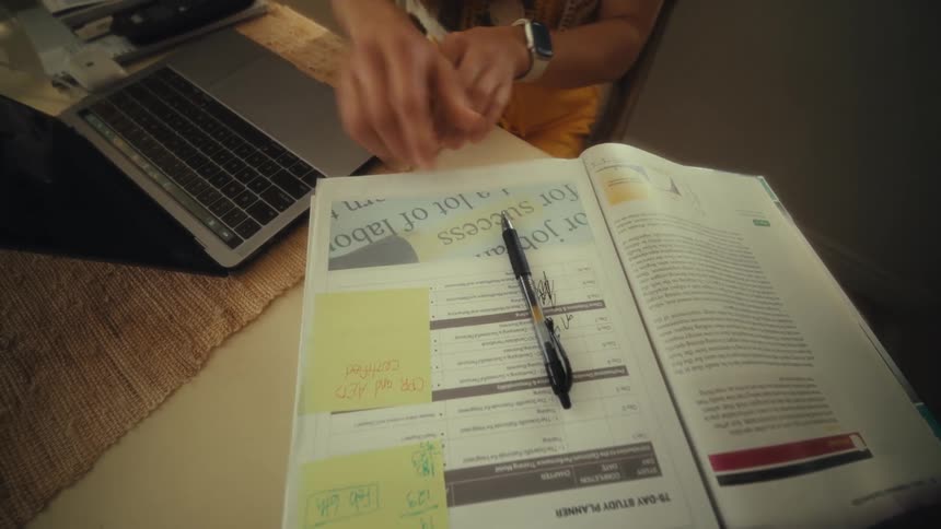 Hand holds a pen and writes in an open book on a table while typing on a laptop