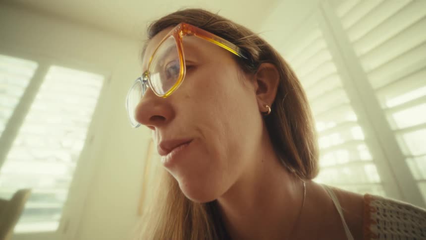 A woman wearing orange glasses and a crochet top sits at a wooden desk writing in a notebook and using a calculator next to a laptop while bright sunlight streams through window shutters in a home office