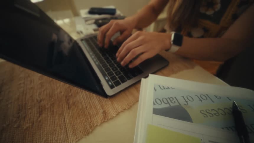 A woman writes a note with a pen and then uses her laptop