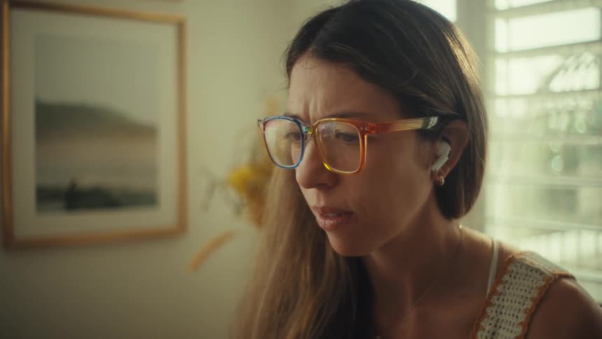 A woman wearing colorful glasses and wireless earbuds speaks during a video call on a laptop while sitting at a wooden table in a bright room with books and a water bottle nearby