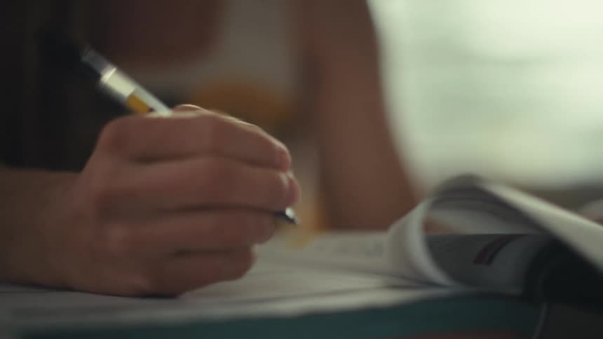 A woman wearing colorful glasses and a crochet top writes in a book with a pen while sitting at a table during the day