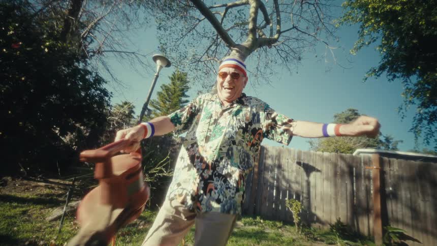 Elderly man wearing a sports headband dances on the lawn while holding a leaf blower