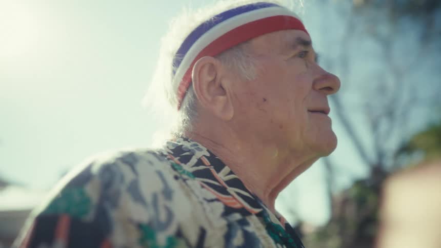 An elderly man wearing a red white and blue headband looks up toward the sky in a sunny outdoor setting he is dressed in a patterned colorful shirt and appears to be breathing deeply or reflecting while bathed in bright sunlight
