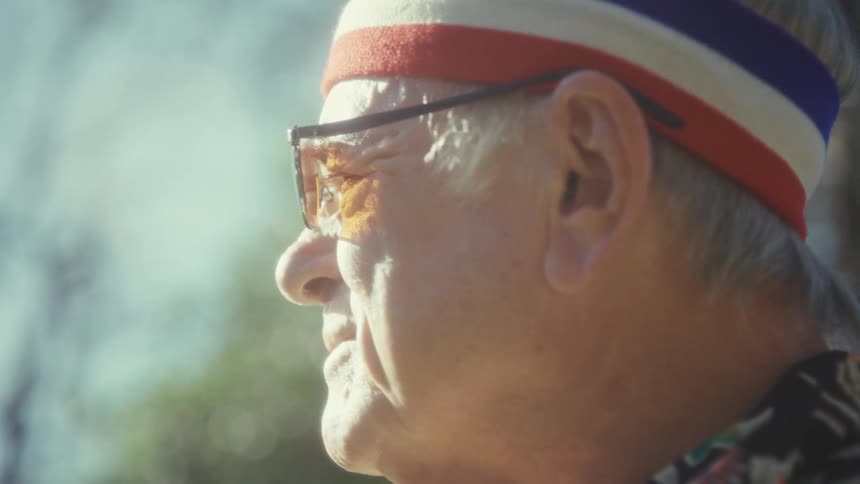 An elderly man wearing glasses and a headband standing outdoors and looking at his surroundings on a sunny day