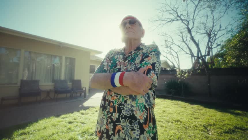 An elderly man wearing a summer shirt wristbands and a headband standing on his backyard lawn under the sun with his arms crossed