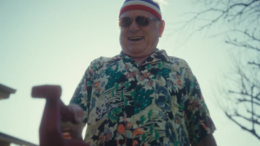 Elderly man wearing a sports headband and sunglasses holds a red object in the backyard