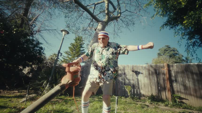 Elderly man wearing a sports headband dances in the backyard while holding a leaf blower