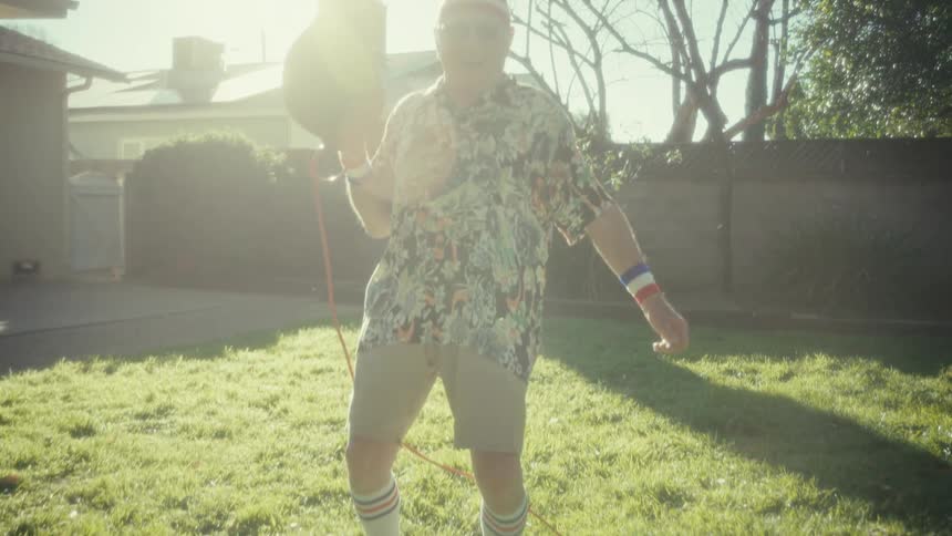 A man in retro workout gear strolls across a bright backyard lawn holding a red leaf blower grinning as he swings it up playfully then the sun flares hard and another hand reaches in to take the blower from him