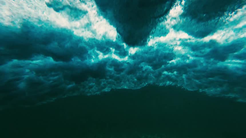A view from under the surface of the ocean shows white foam and bubbles from a breaking wave above a sandy sea floor with sunlight patterns