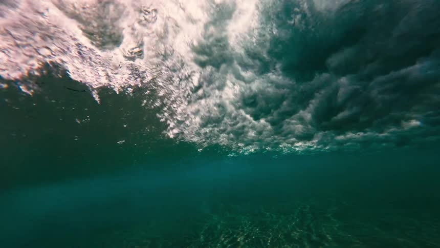 Sunlight ripples across the sandy ocean floor before the camera rises to meet a wave crashing overheadthe underwater view captures the transformation of the clear surface into a chaotic explosion of white foam and bubbles
