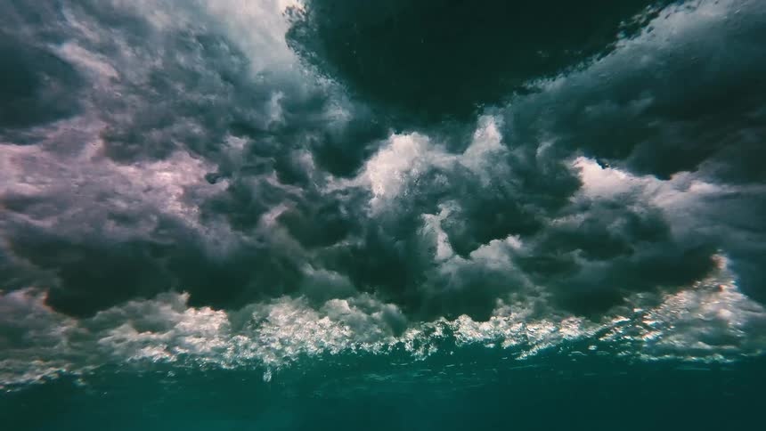 A view from under the water shows a thick layer of white foam and dark swirling bubbles from a crashing wave above a clear blue sea
