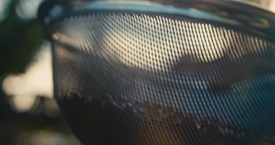 A metal colander containing dark coffee beans is shaken and tossed upward in a outdoor setting with a blurred background of trees and soft sunlight