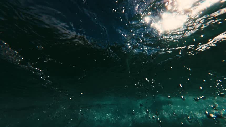 A view from under the water shows a thick layer of white foam and dark swirling bubbles from a crashing wave above a clear blue sea