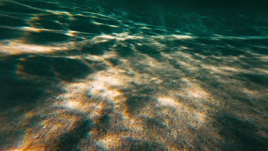 Sunlight filters through the surface of the ocean creating dancing patterns of light and shadow across a sandy seabedthe camera glides slowly forward over the underwater terrain capturing the rhythmic movement of turquoise water