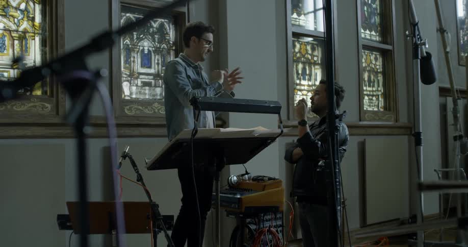 Two men stand at a lectern talking in a building with stained glass and recording equipment