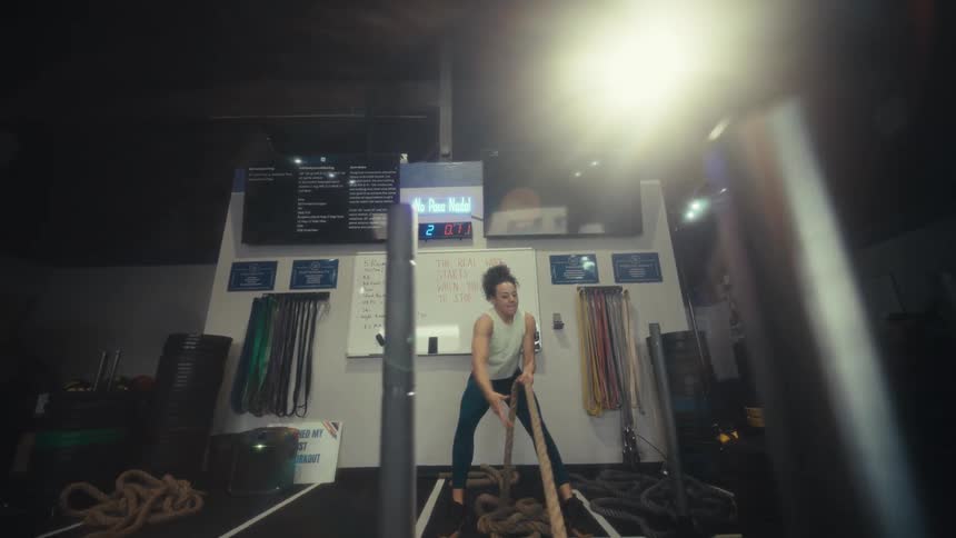 A woman wearing a sleeveless top and dark pants pulls a heavy rope toward her while exercising in a gym with weights and fitness equipment in the background