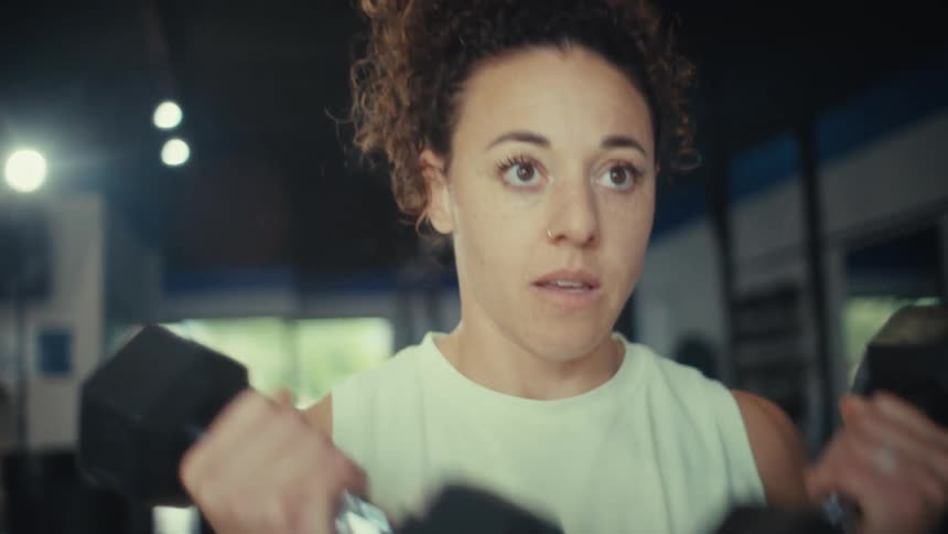 A woman with curly hair performs bicep curls using black dumbbells inside a gym
