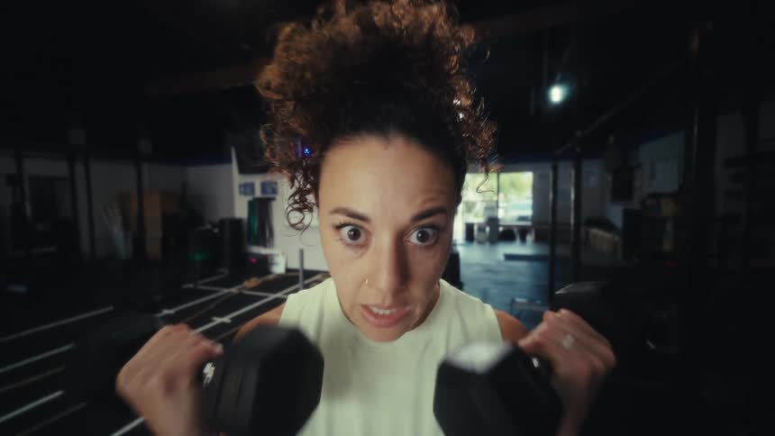 A woman wearing a white sleeveless top and dark leggings performs bicep curls with dumbbells while looking directly at the camera in a gym setting