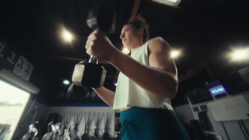 A woman in a white crop top and teal leggings performs bicep curls with heavy dumbbells inside a spacious gym