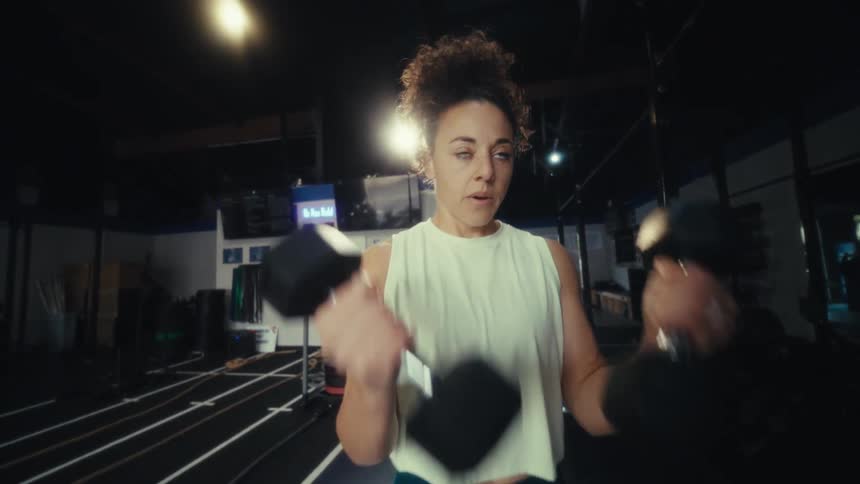 A woman is performing dumbbell bicep curls inside a gym settingthe gym features dark flooring with several white parallel lines and a sled track in the background