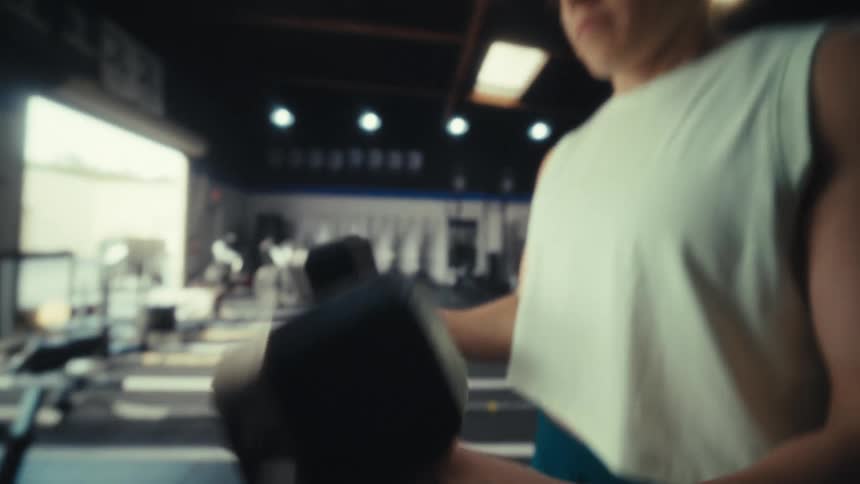 A woman in a white tanktop performs bicep curls with heavy dumbbells inside a sunlit gym