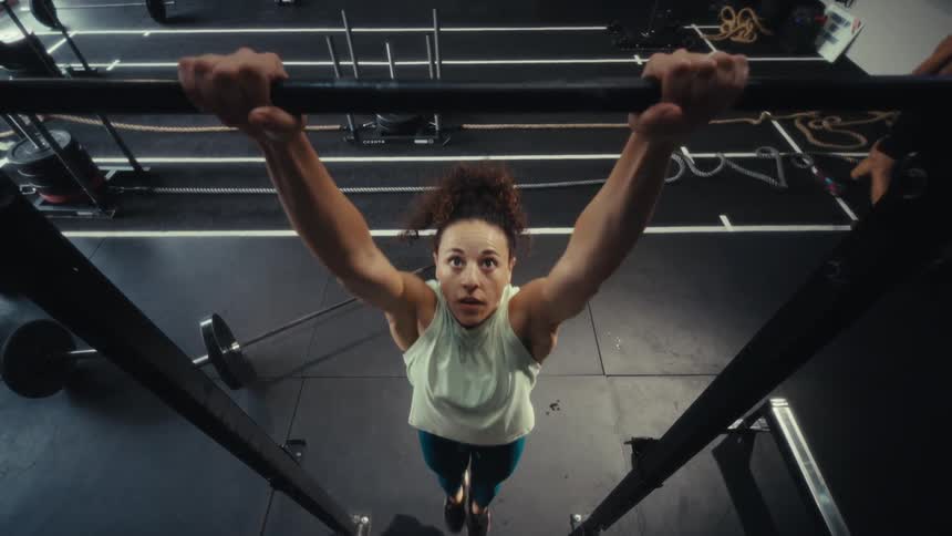 A woman with curly hair wearing a light green tank top and blue leggings performs pull ups on a black metal bar in a gym with weights and training equipment in the background