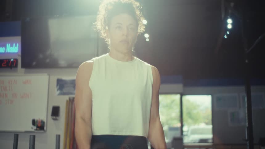 A woman with curly hair stands in a gym performing bicep curls with a pair of dumbbells her shadow is cast prominently on the floor as she maintains a focused expression under the bright overhead lights