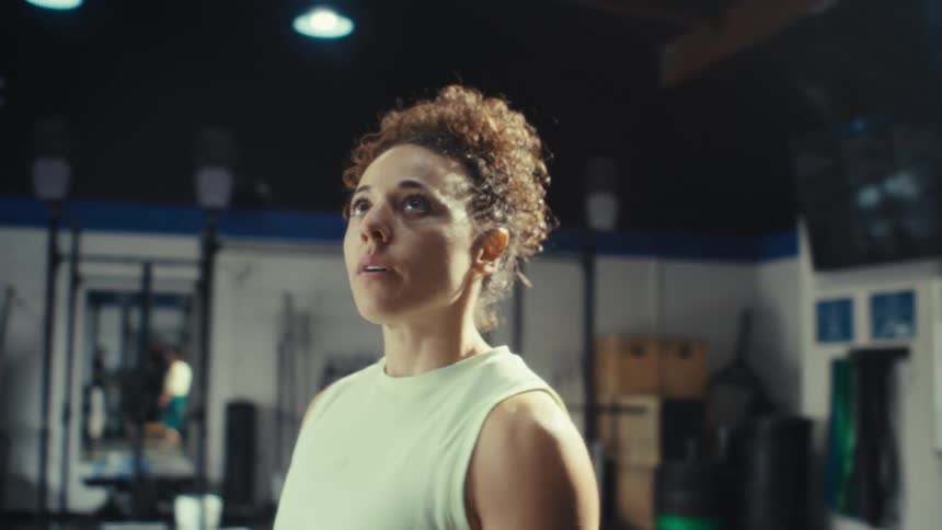 A woman with curly hair stands in a gym and looks upward while breathing heavily during a workout