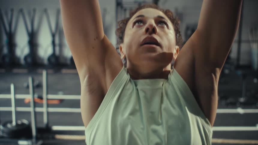 A woman with curly hair prepares herself and begins performing pullups at a gym the camera focuses on her concentrated expression as she grips the bar and pulls her chin above it