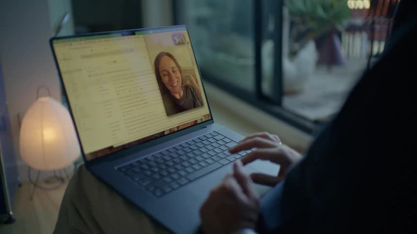 A view from behind a man shows him having a video call on a laptop with a woman who smiles and waves while a paper lamp glows in a dimly lit room with a dark window in the background