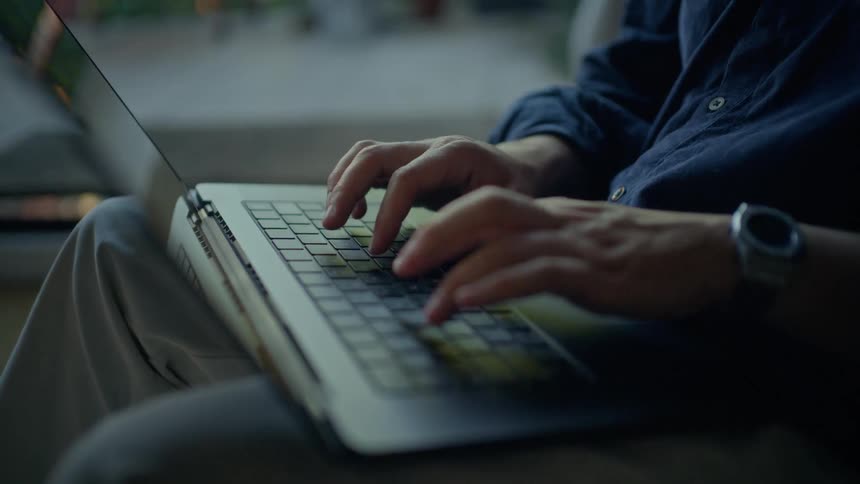 A close view of a man wearing a blue shirt and a watch typing on a laptop keyboard while sitting outdoors in the evening