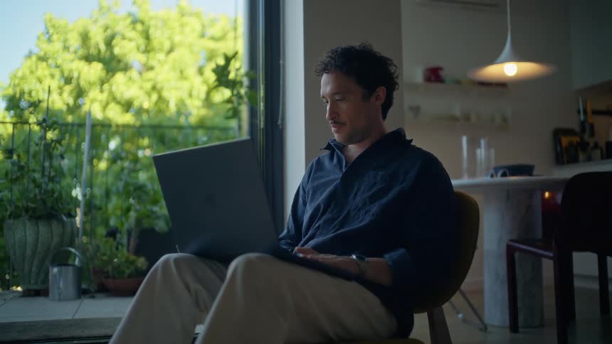 Man sitting on a chair works on a laptop near a window with a view of plants
