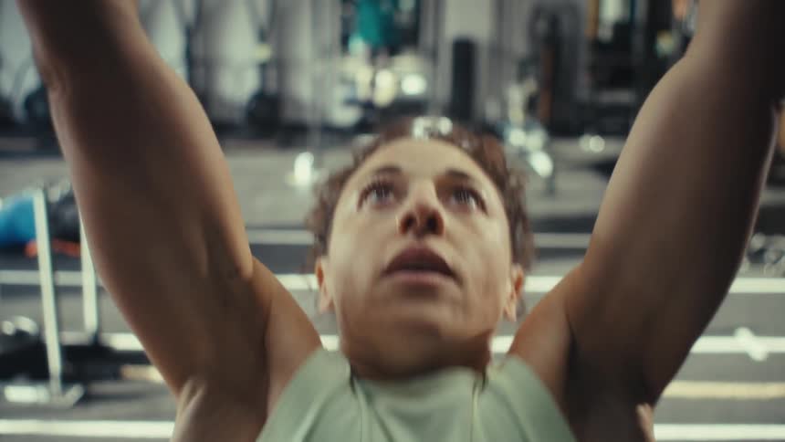 A woman with curly hair wearing a white sleeveless top and blue leggings performs pull ups on a black metal bar in a gym with weights and training equipment in the background