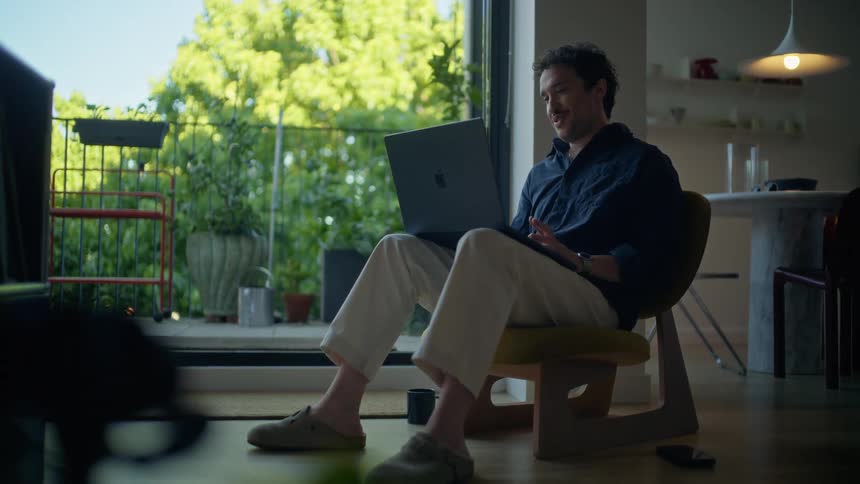 A young man sitting on a stool in his corridor with a laptop on his lap having a video call with green vegetation visible outside the building at midday
