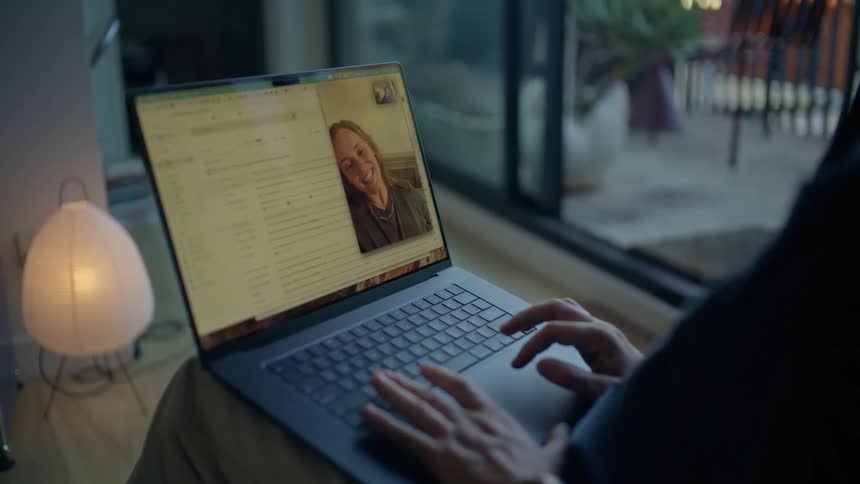 A person is seen using a laptop to engage in a video call with a smiling woman the laptop screen displays a split view of the video chat alongside what appears to be an email or document interface