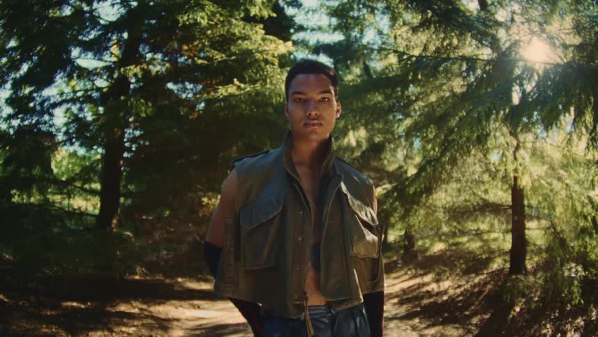 A young man stands in a sunlit forest looking directly at the camera while wearing a cropped utility vest and blue jeans as light filters through the dense evergreen branches and creates a bright glow behind his head in the woods