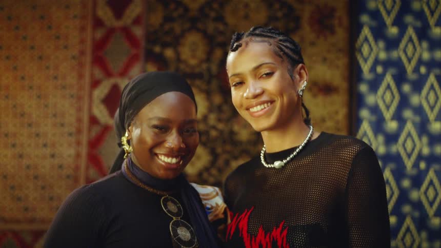 Two women smile and look toward the camera while standing together in front of a background of hanging textiles with various patterns and colors