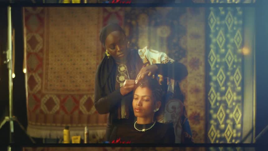 A woman wearing a dark patterned top and large gold earrings stands behind another woman and adjusts her hair as they pose together in a room with various colorful hanging rugs