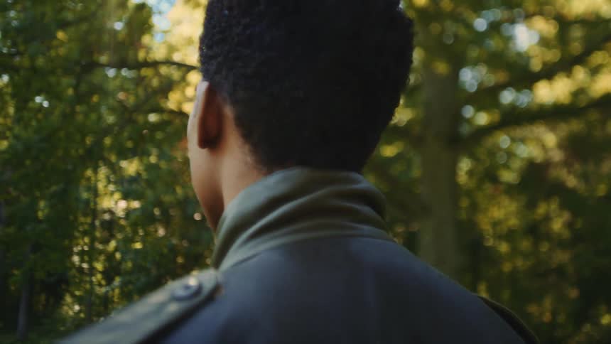 A view from behind a man with short dark hair wearing a green jacket as he looks toward lush green trees in a sunlit forest