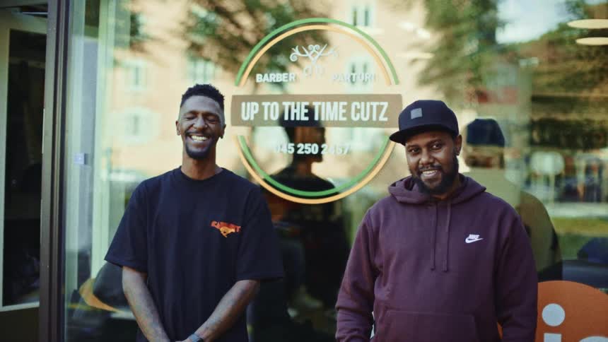 Two men pose smiling in front of a barbershop with a sign on the window