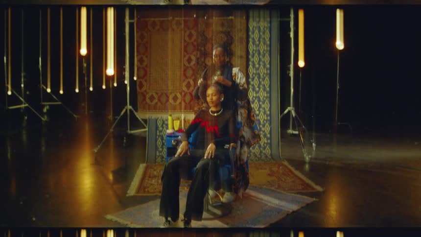 A person stands behind another person seated in a chair and styles their hair in a room with several rugs hanging in the background and warm lights hanging from the ceiling