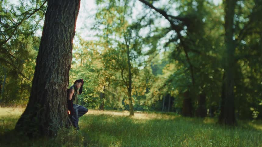 A man with dark hair leans against a large tree trunk in a sunny forest with green grass and many trees in the background