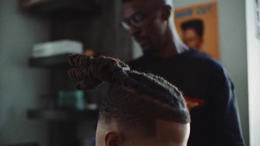 A side view shows a barber wearing glasses and a dark shirt using clippers to trim the hair of a man with a beard and long dreadlocks tied on top of his head inside a shop