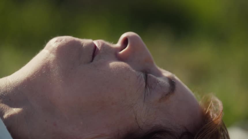 Closeup of a persons face with closed eyes illuminated by natural light against a blurred green background