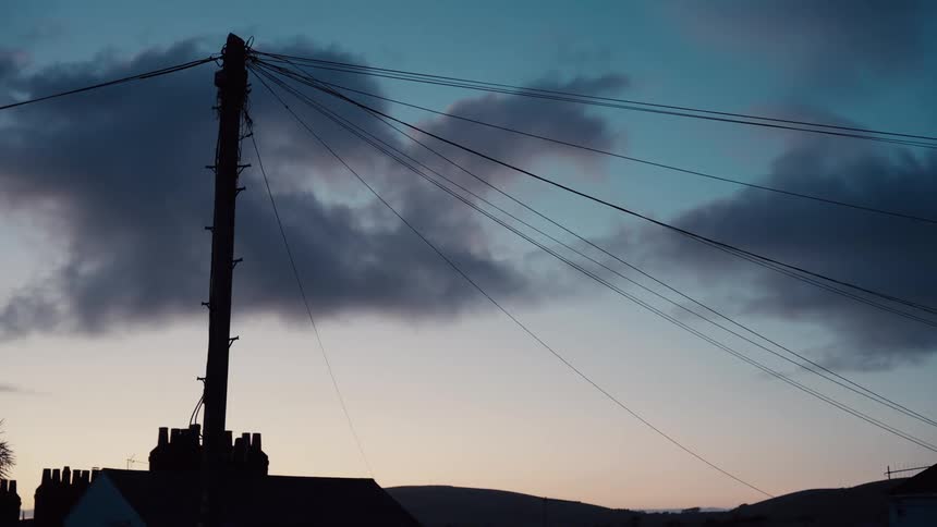 A tall telegraph pole stands silhouetted against a fading blue and orange sky while power lines stretch over the dark outlines of houses and distant hills