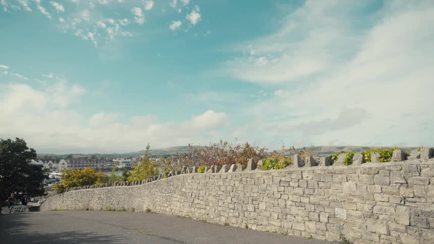 Two people walking uphill on a quiet paved road with brick fencing along the sides and a city skyline visible in the distance at midday