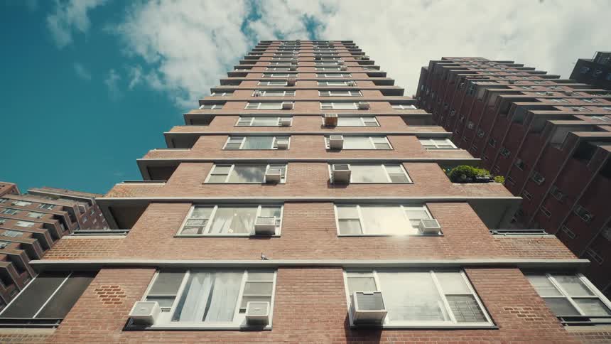 A low angle shot looks up at a tall brick apartment building featuring several rows of windows with installed air conditioning units under a blue sky with light clouds